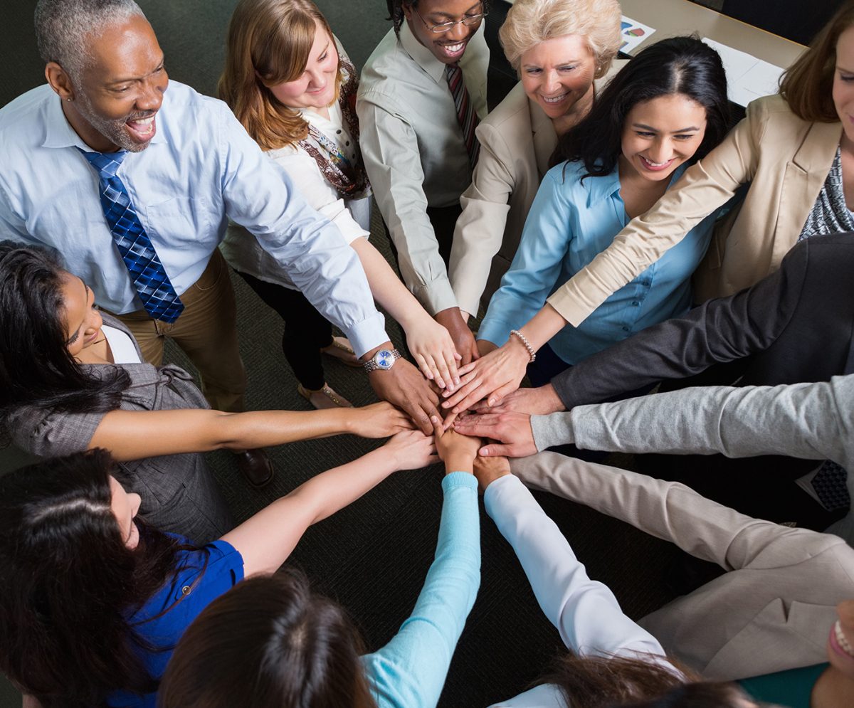 Diverse group of seniors, mature adults, mid adults and young adults are part of team in professional office. They are standing in a circle with their hands  together in middle to celebrate success or teamwork. They are all wearing business casual clothing and smiling.